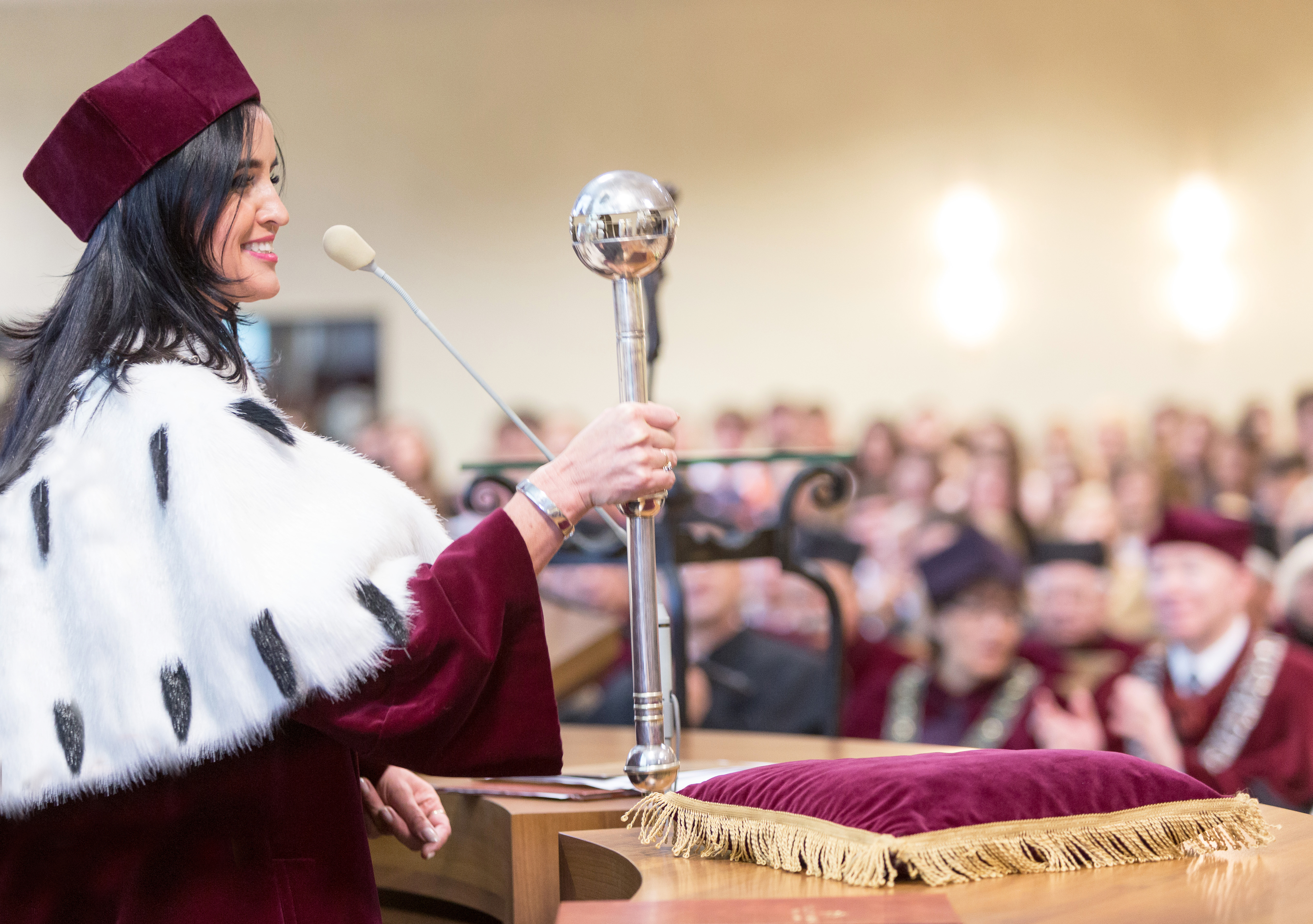 Kobieta w akademickim stroju prezentuje insygnium podczas uroczystości. W tle widać zgromadzonych gości.
