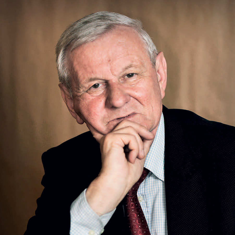 Mężczyzna w garniturze z ręką przy brodzie, patrzący w kierunku kamery. Tło w neutralnym kolorze.