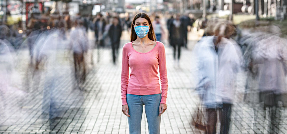 Kobieta w różowej bluzce i niebieskiej masce, stojąca na chodniku wśród tłumu.