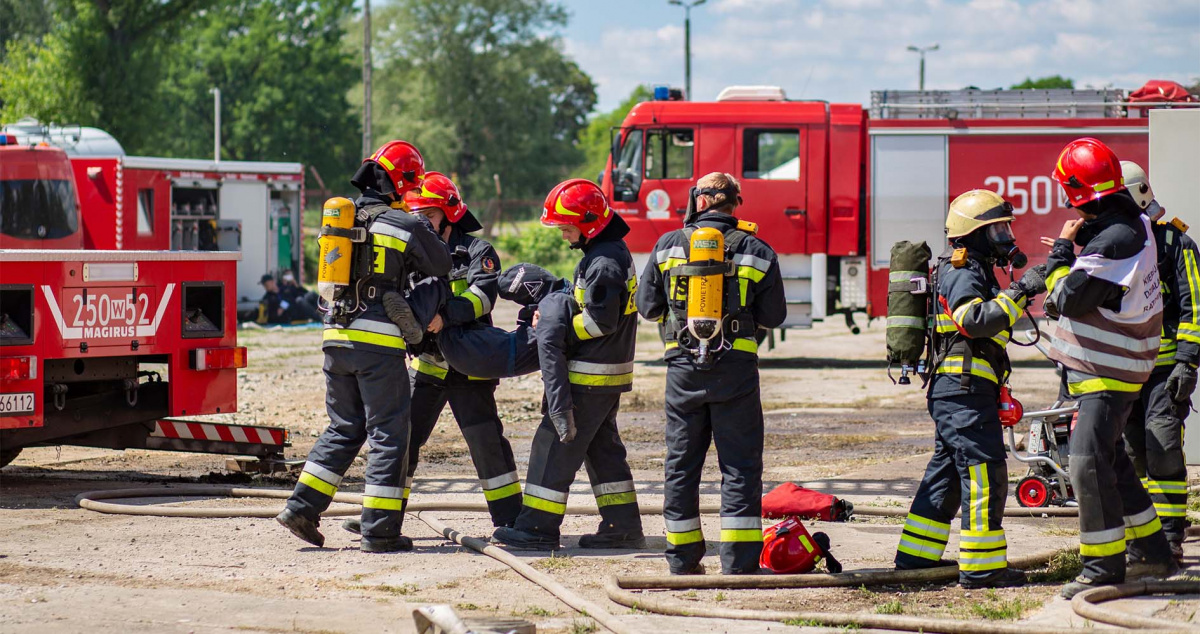 Grupa strażaków w akcji, z wozem strażackim w tle.