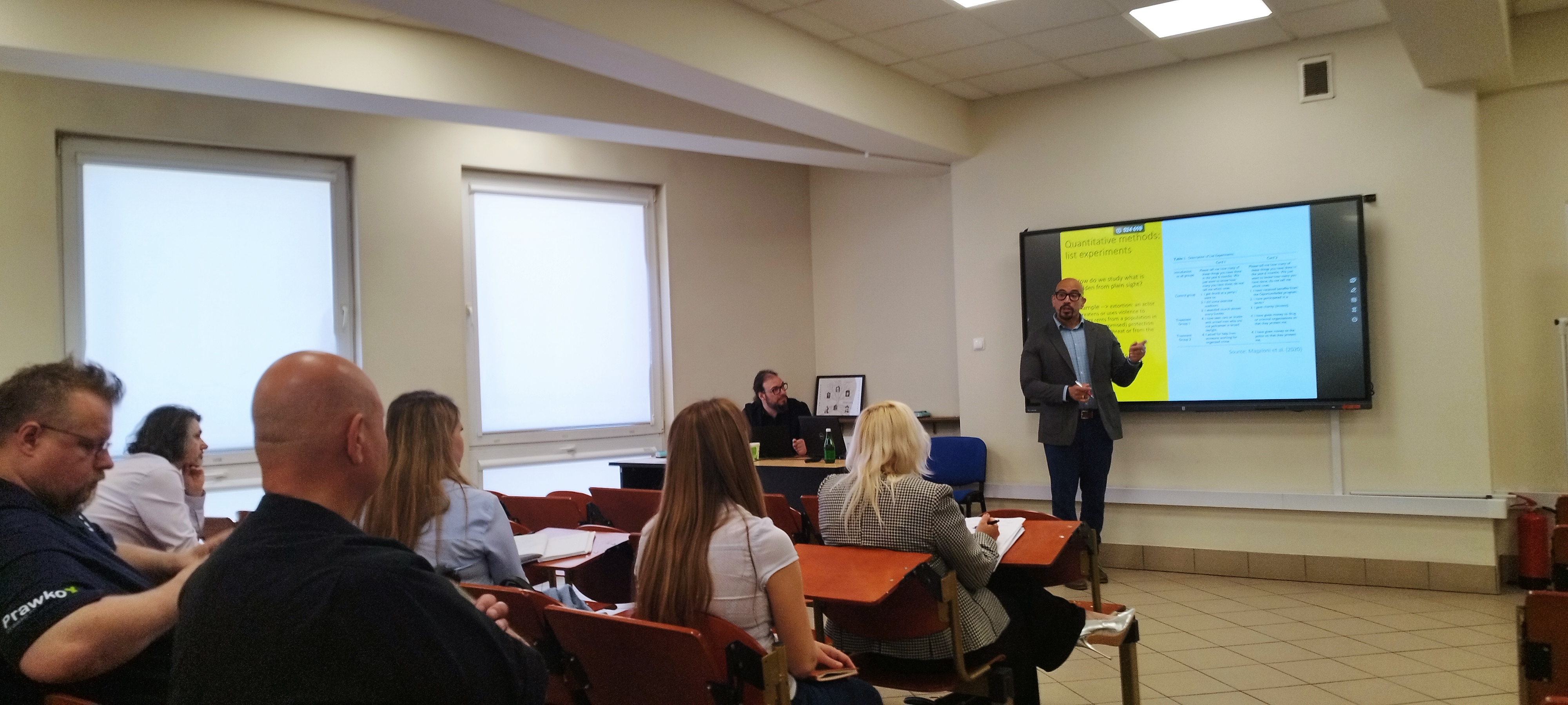 Prezentacja w sali wykładowej z uczestnikami siedzącymi w rzędach, w tle widoczny ekran z materiałami.