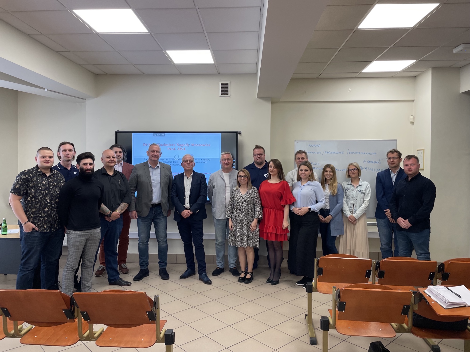 Grupa osób pozująca do zdjęcia w sali wykładowej, z prezentacją w tle.