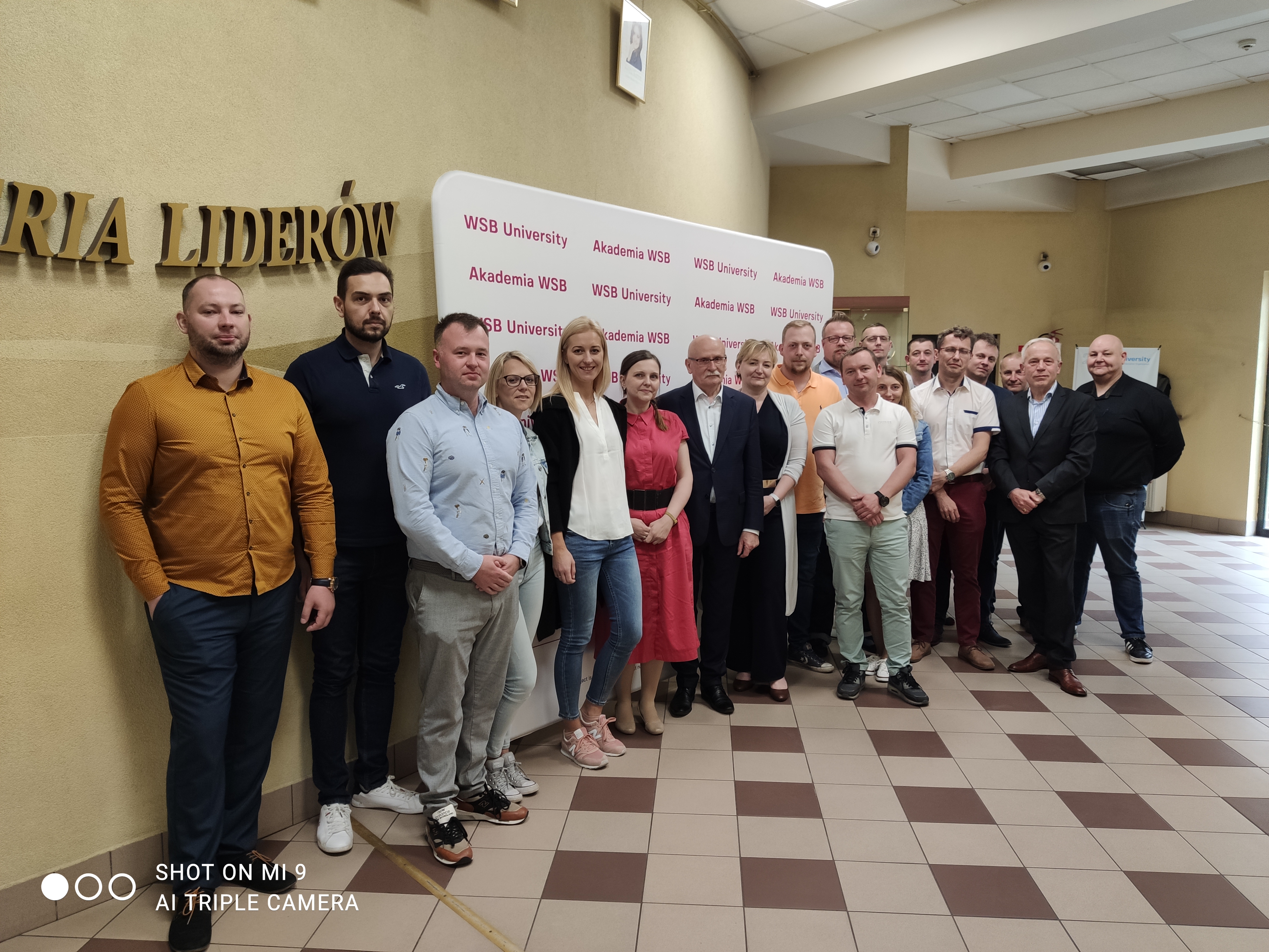 Grupa osób stoi w pomieszczeniu z logo Akademii WSB na tle.
