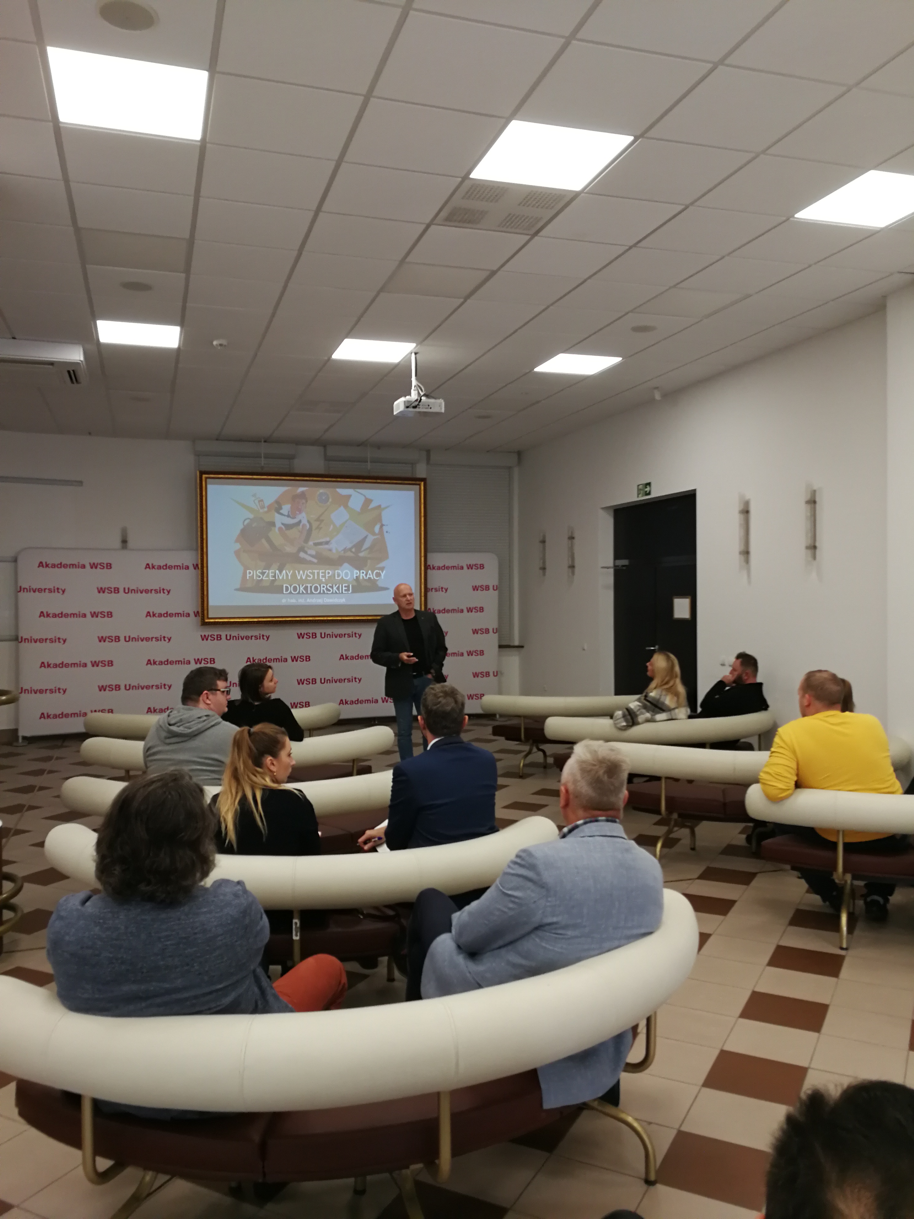 Spotkanie w Akademii WSB, podczas którego omawiane są zasady tworzenia prac doktorskich. Uczestnicy siedzą w okręgu, a na ekranie wyświetlana jest prezentacja.