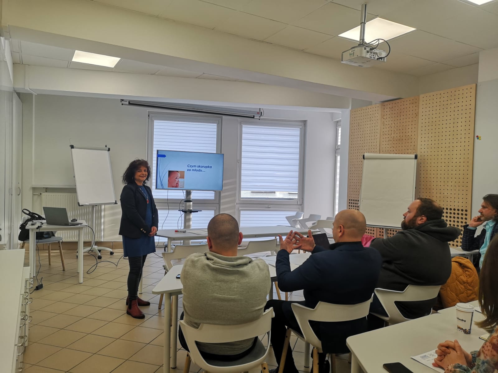 Prezentacja odbywająca się w jasnym pomieszczeniu, z uczestnikami siedzącymi przy stołach, a prowadząca stoi obok wyświetlacza.