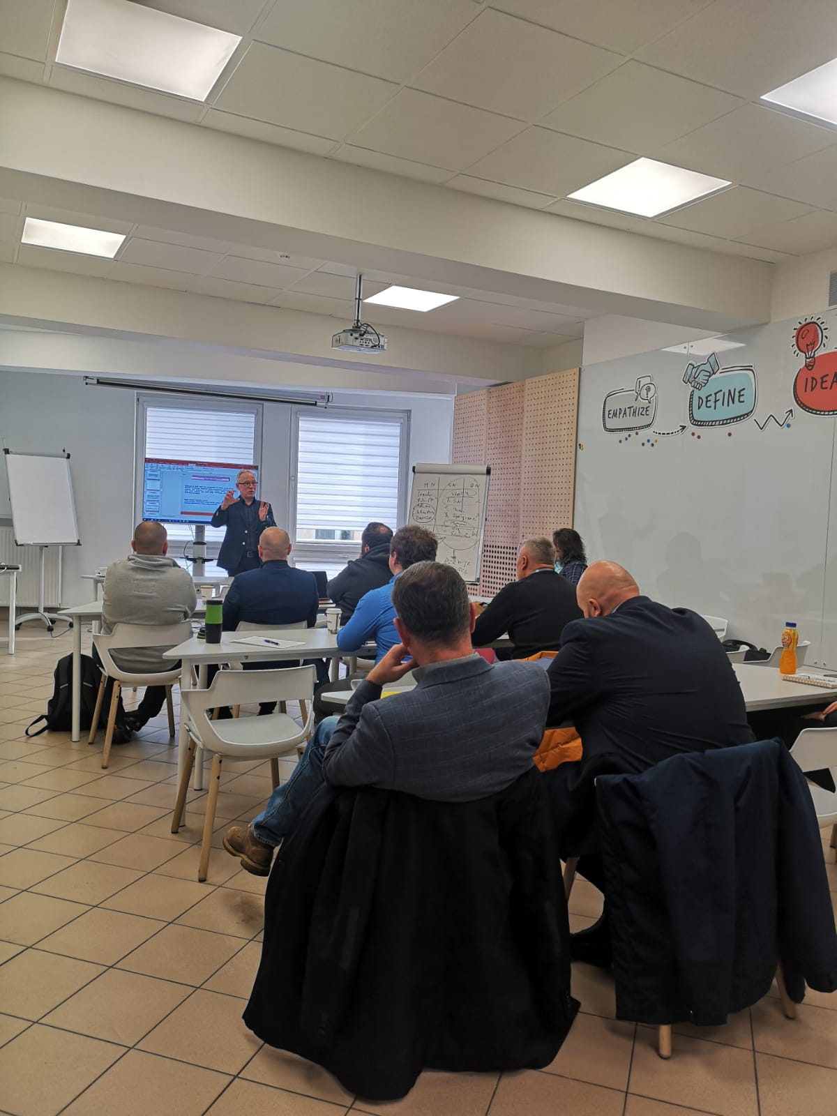 Spotkanie w sali konferencyjnej, gdzie prowadzący prezentuje informacje dla grupy uczestników.