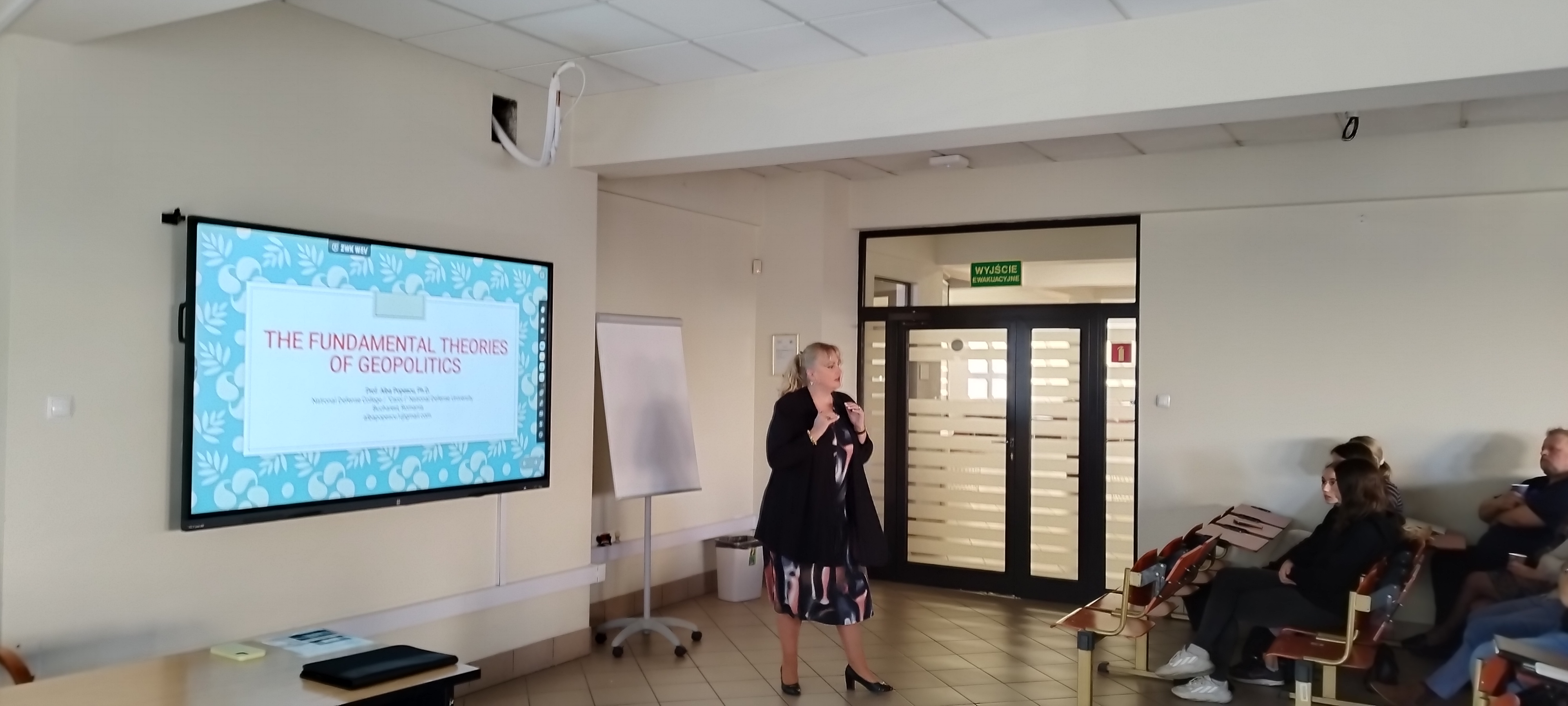 Prezentacja na temat podstawowych teorii geopolityki. W tle widoczna publiczność i ekran z prezentacją.